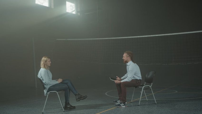 A man and a woman engage in a therapy session in a smoky indoor setting, sitting apart in a large room.